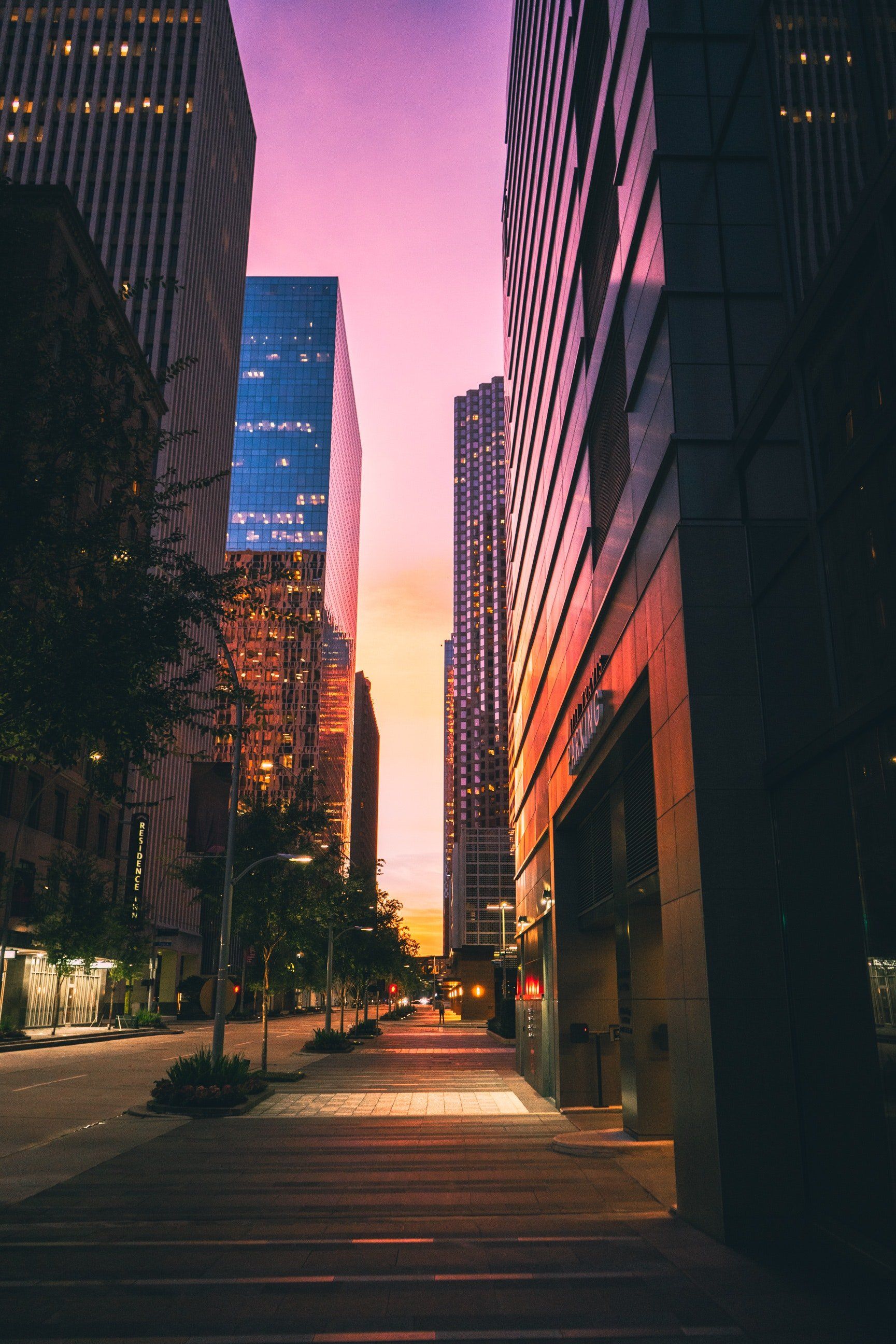 Skyscrapers line a city street at sunset, under a purple and orange sky.